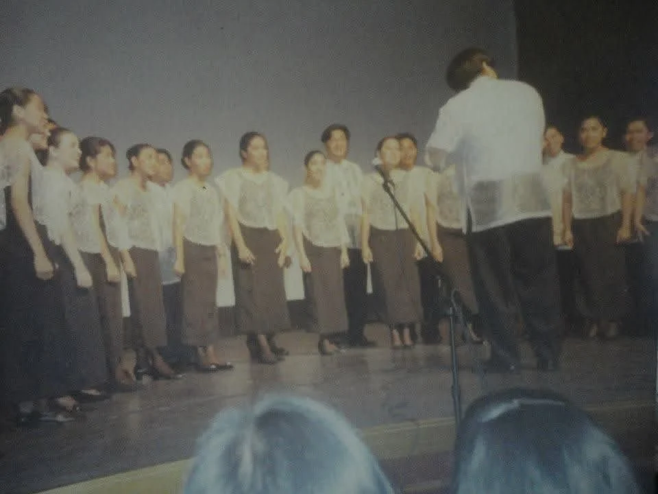 A choir in barong and traditional dress on a stage, singing to a conductor whose back is to the camera